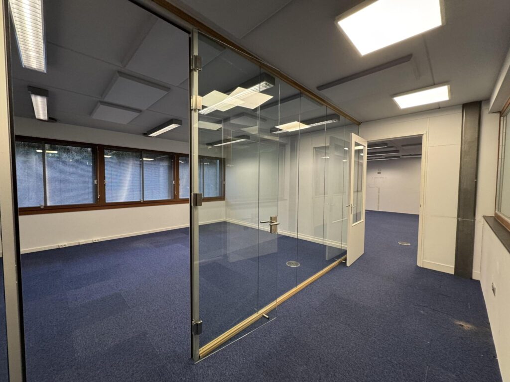 Modern office space at De Ruijterkade with glass-walled meeting room and blue carpet flooring.